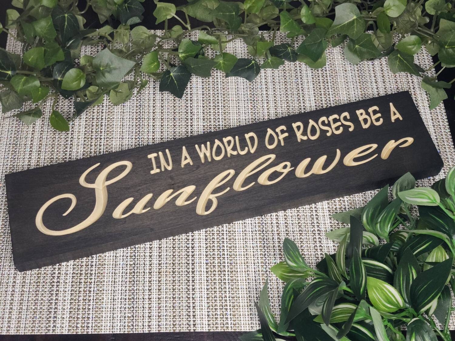 "In a World Full of Roses, Be a Sunflower" wooden sign displayed in a bright, cozy living room.