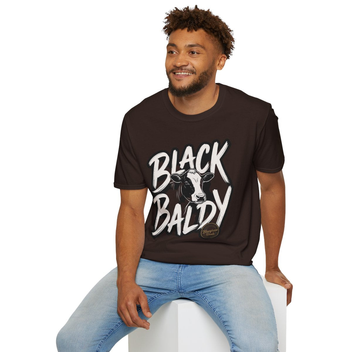 A man with curly hair seated on a white block, smiling, wearing a dark brown Cattle Shirt - Black Baldy