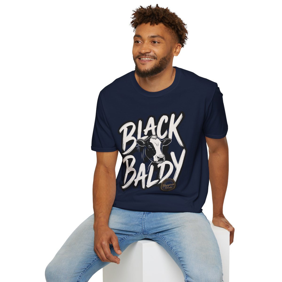 A man sitting on a white block, wearing a navy blue Cattle Shirt - Black Baldy, with the full graphic visible