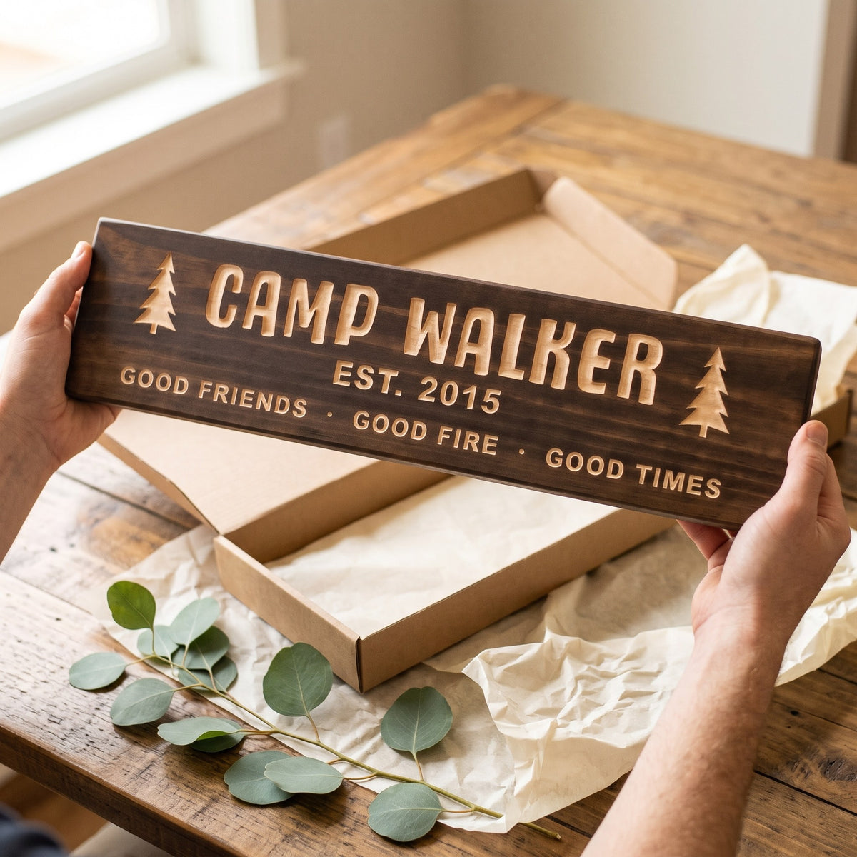 Hands holding a personalized wood sign that reads Camp Walker over a kraft gift box with tissue paper and twine