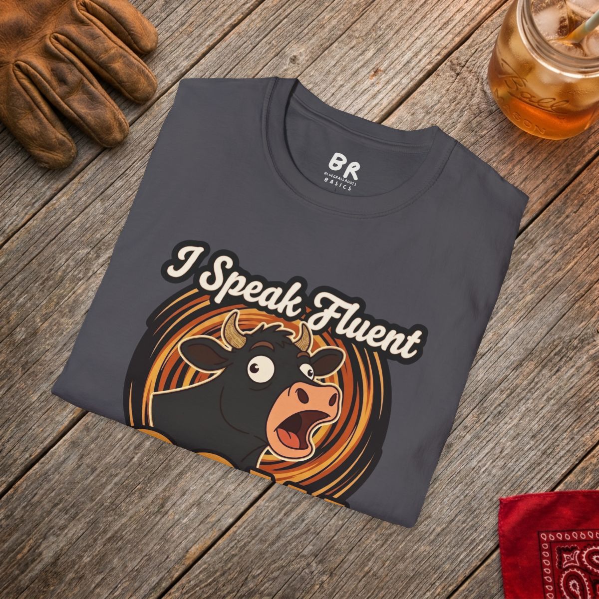 A charcoal gray Farmer Shirt - I Speak Fluent Cow, folded on a rustic wooden table with a work glove and iced tea
