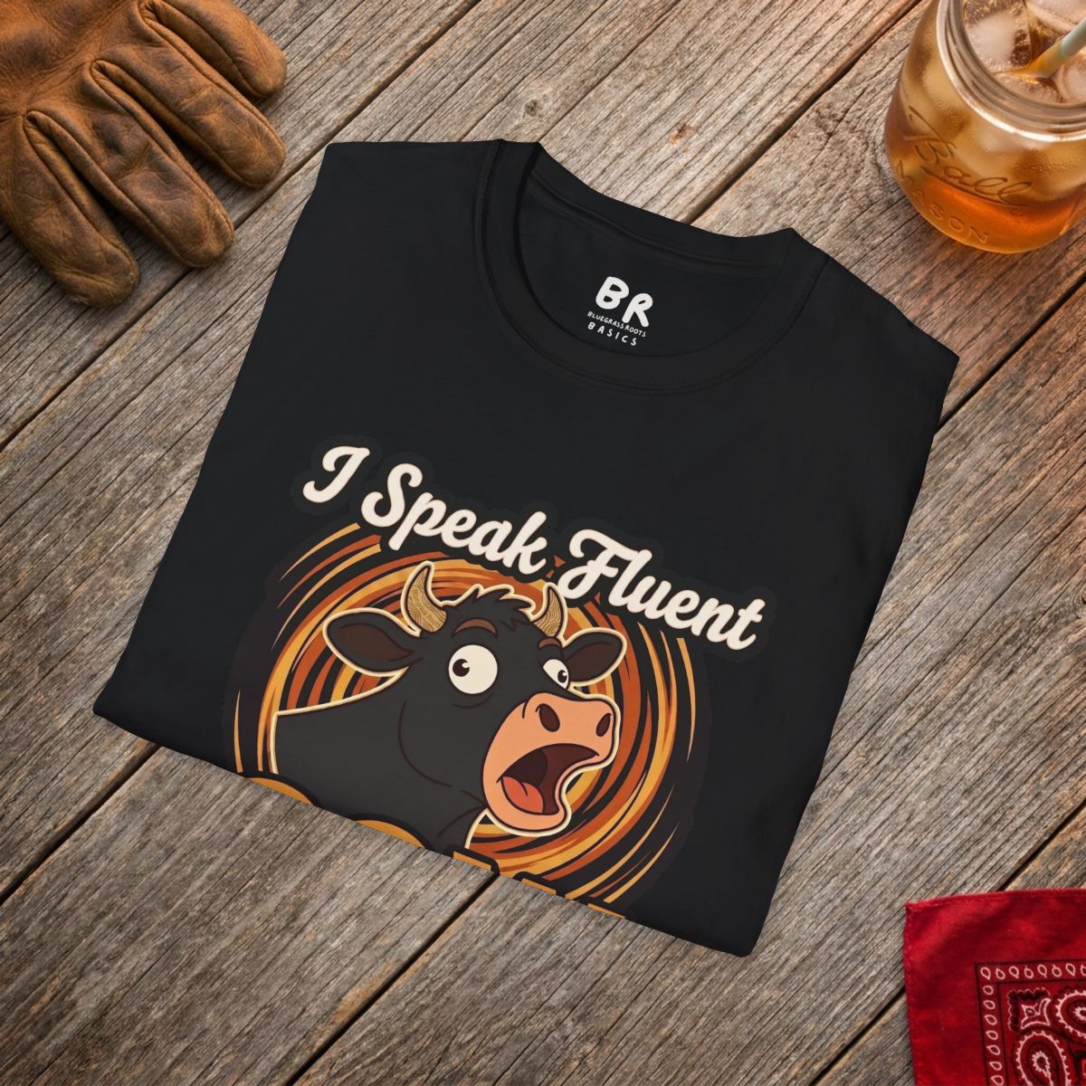 A black Farmer Shirt - I Speak Fluent Cow, folded on a rustic wooden table with a work glove and iced tea