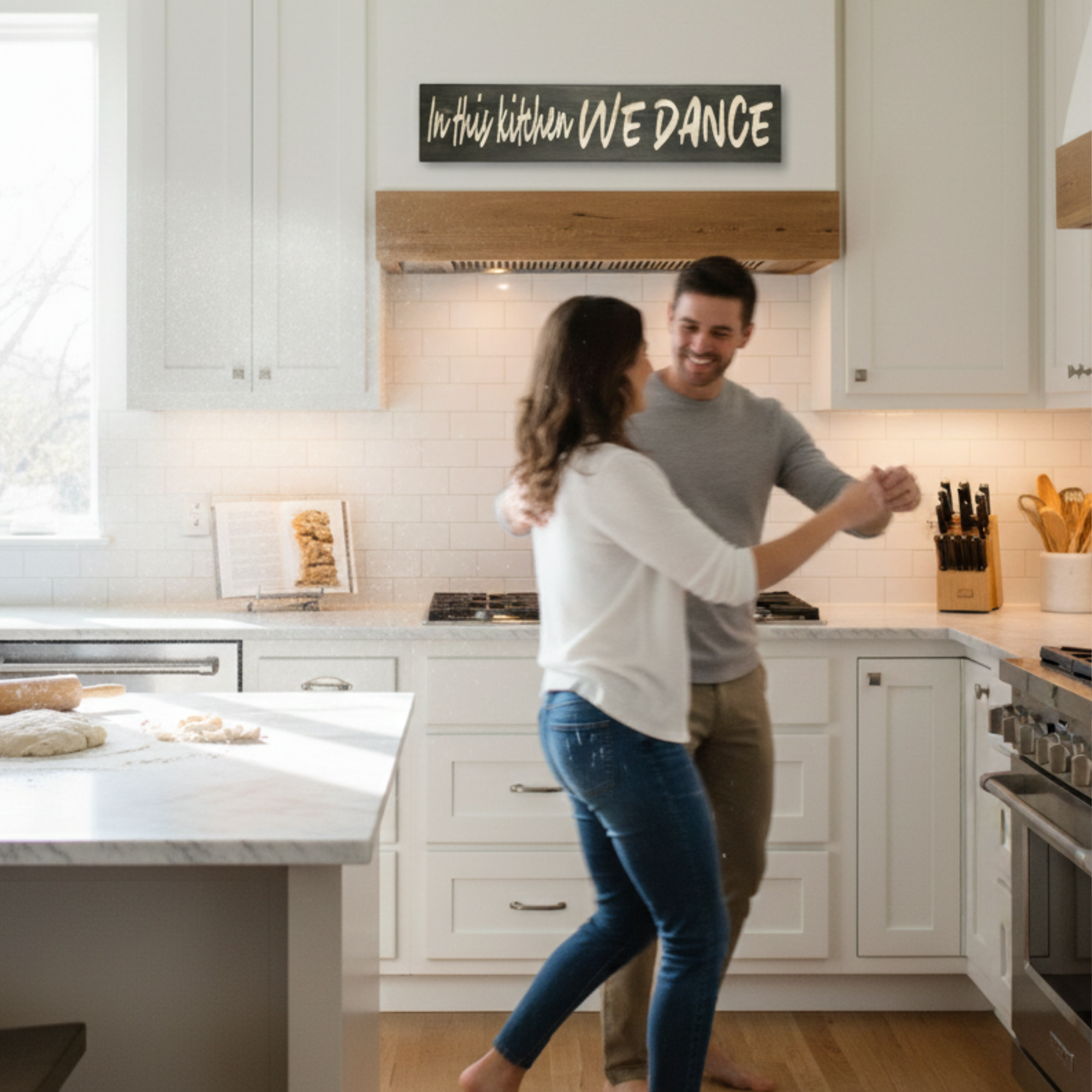 A happy couple dancing in a modern farmhouse kitchen with the "In This Kitchen We Dance" carved wood sign displayed as a focal point above the stove.