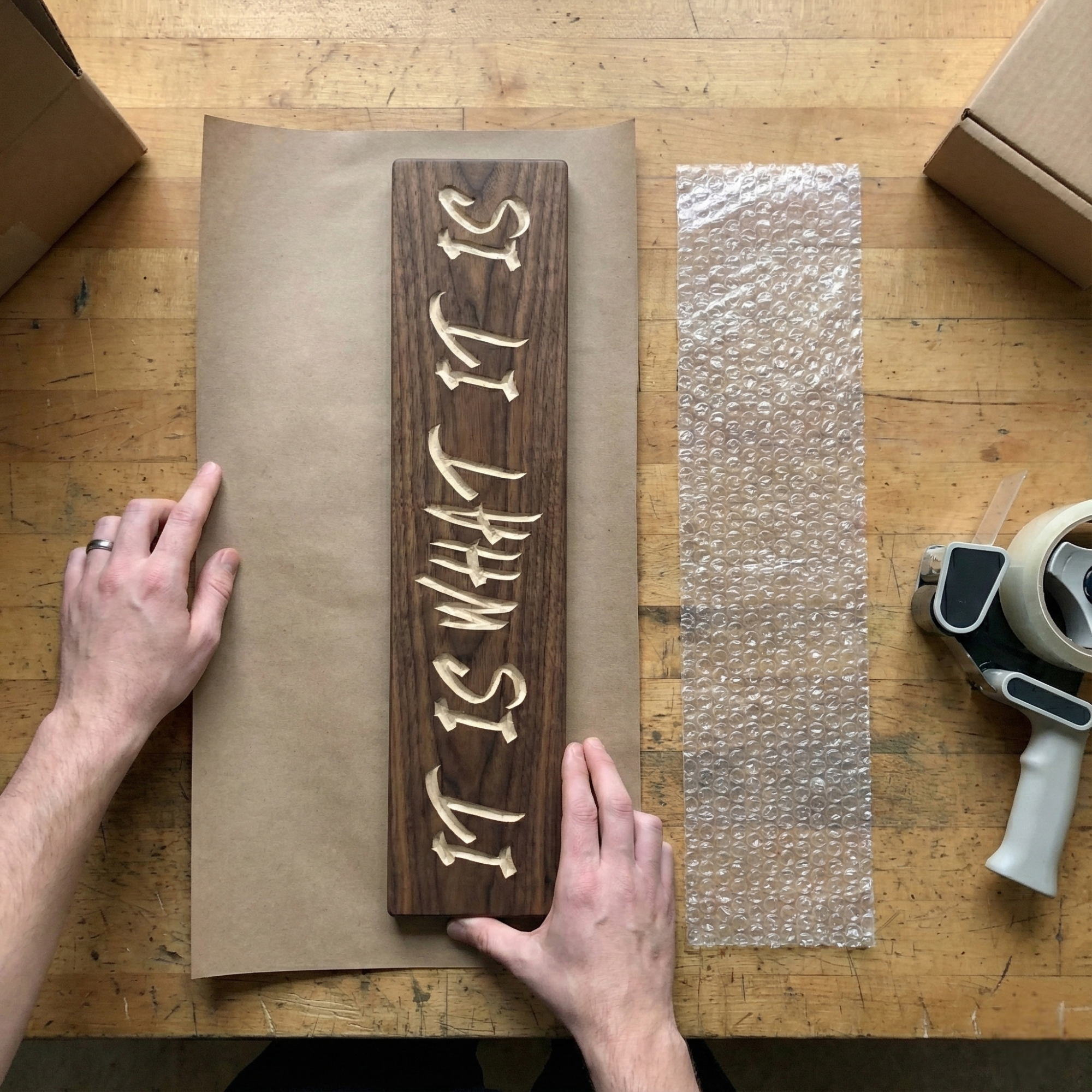 Overhead view of hands preparing a handcrafted wooden IT IS WHAT IT IS sign for shipping with paper and bubble wrap on a workbench.