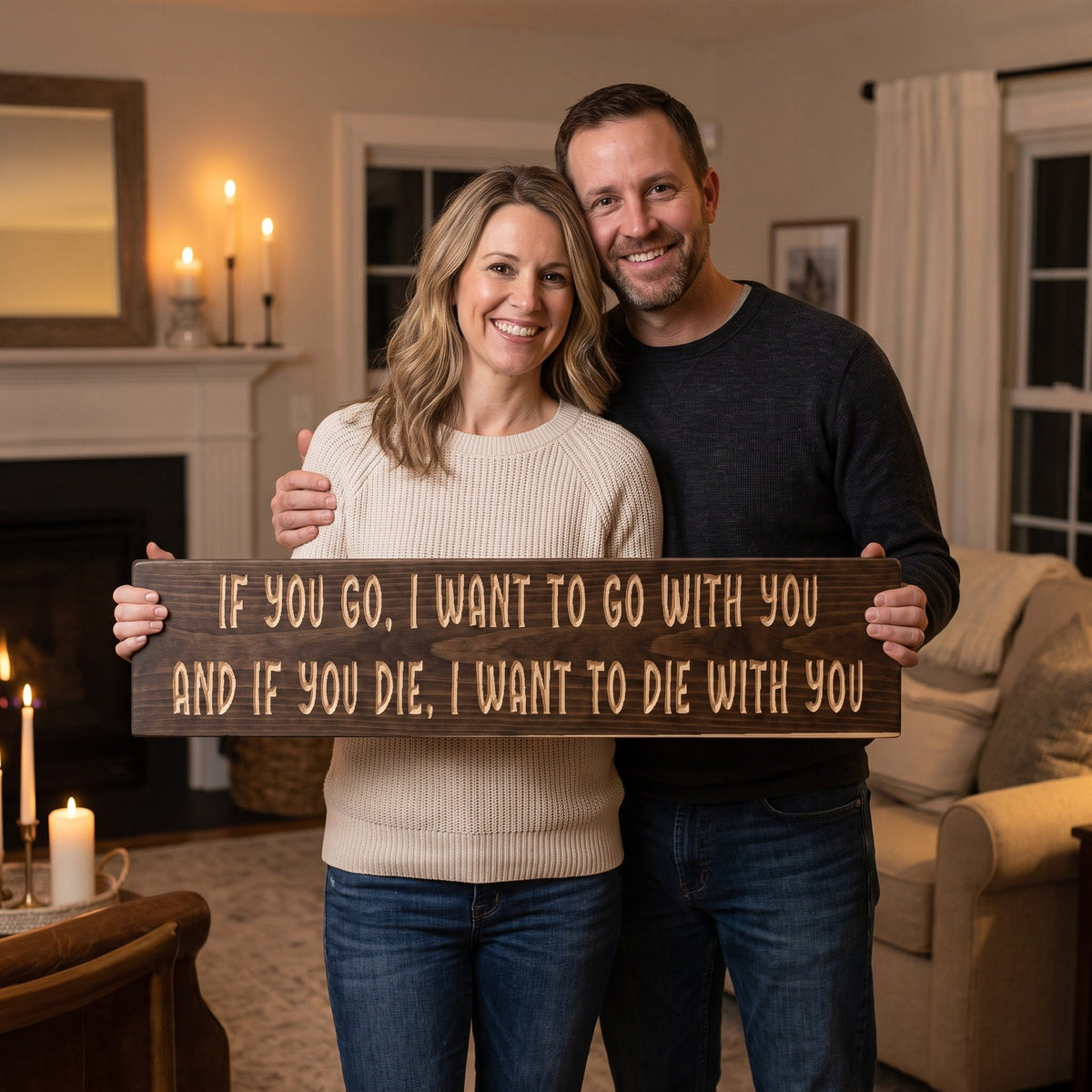 A smiling woman holds the If You Go, I Want to Go With You sign while her husband hugs her in their living room
