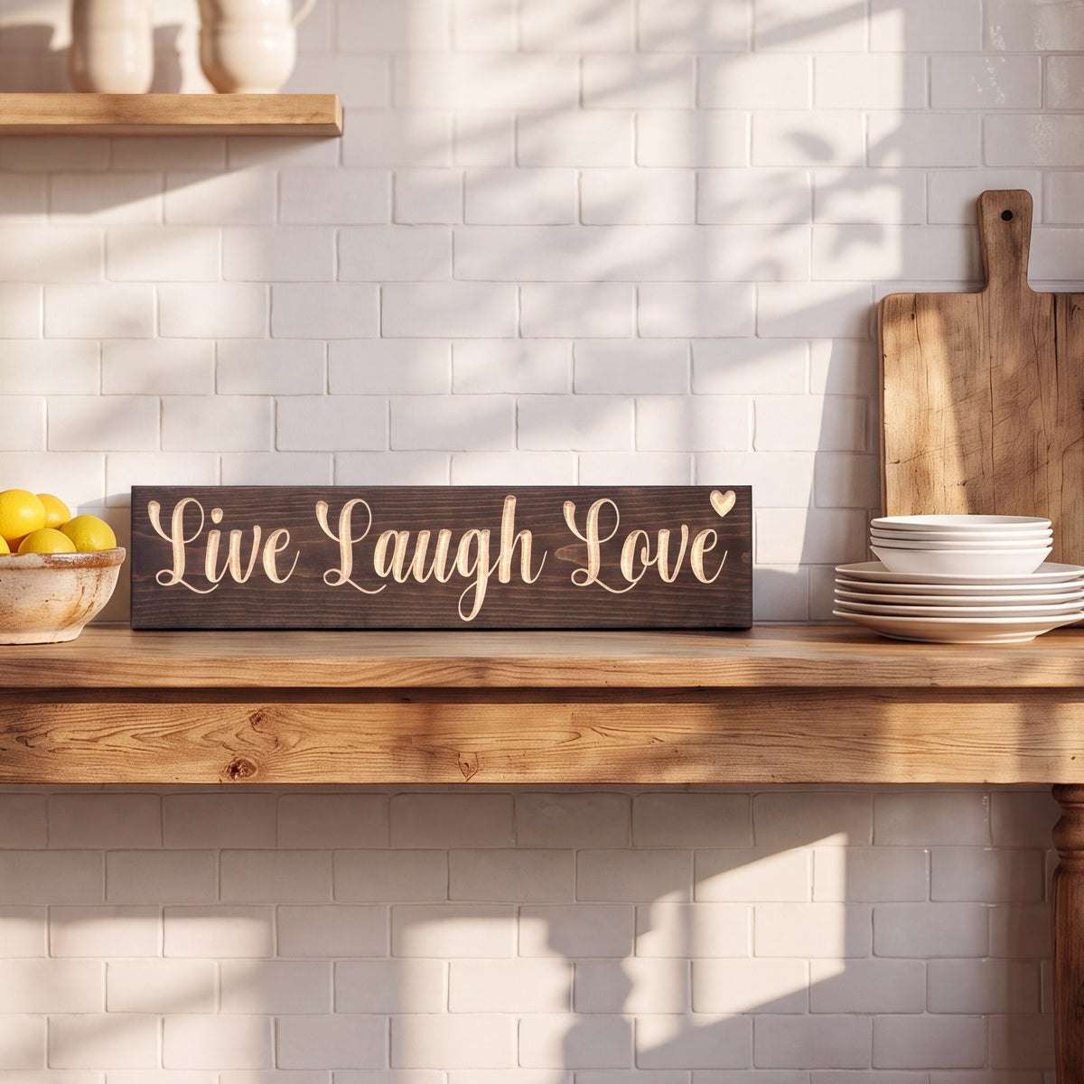 The Live Laugh Love sign leaning on a kitchen console table between a bowl of lemons and a stack of white plates