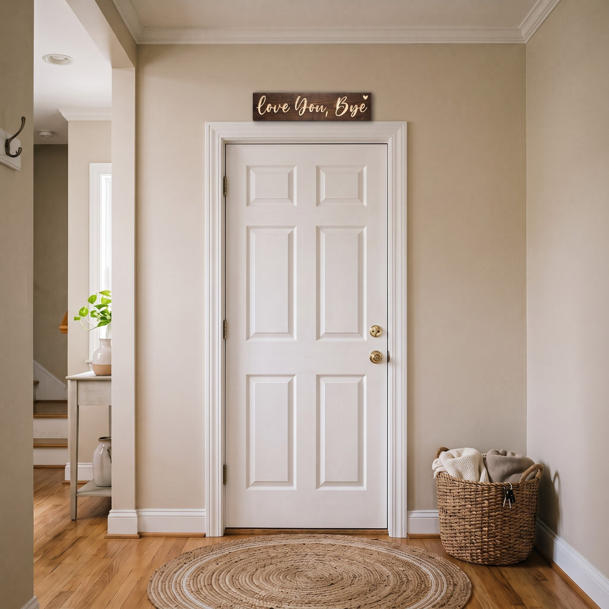 The Love You, Bye sign hanging above the inside of a white front door in an entryway, a basket and a round jute rug below