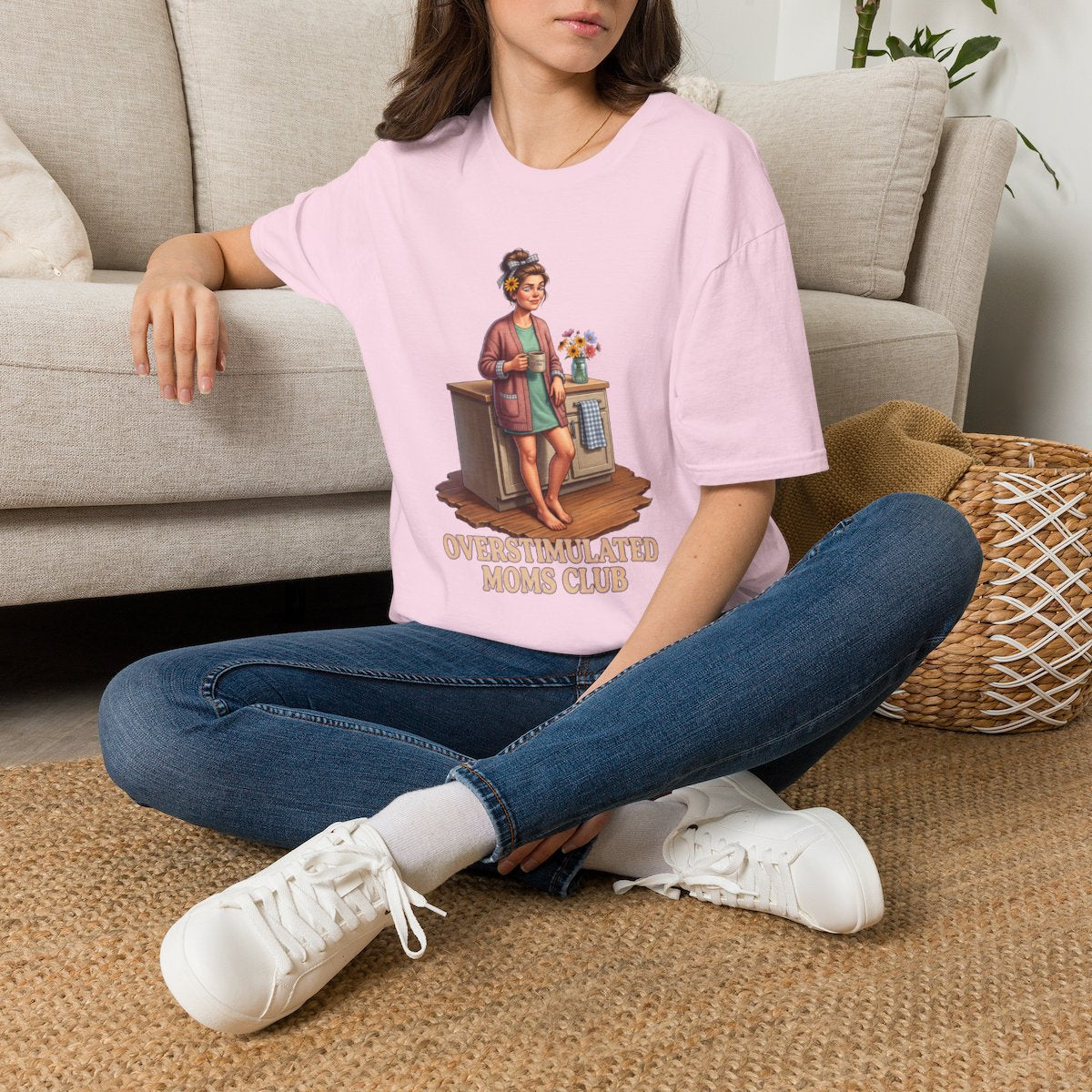 A woman sits on a rug, wearing a pink Overstimulated Shirt - Overstimulated Moms Club, with a couch in the background from Bluegrass Gifts