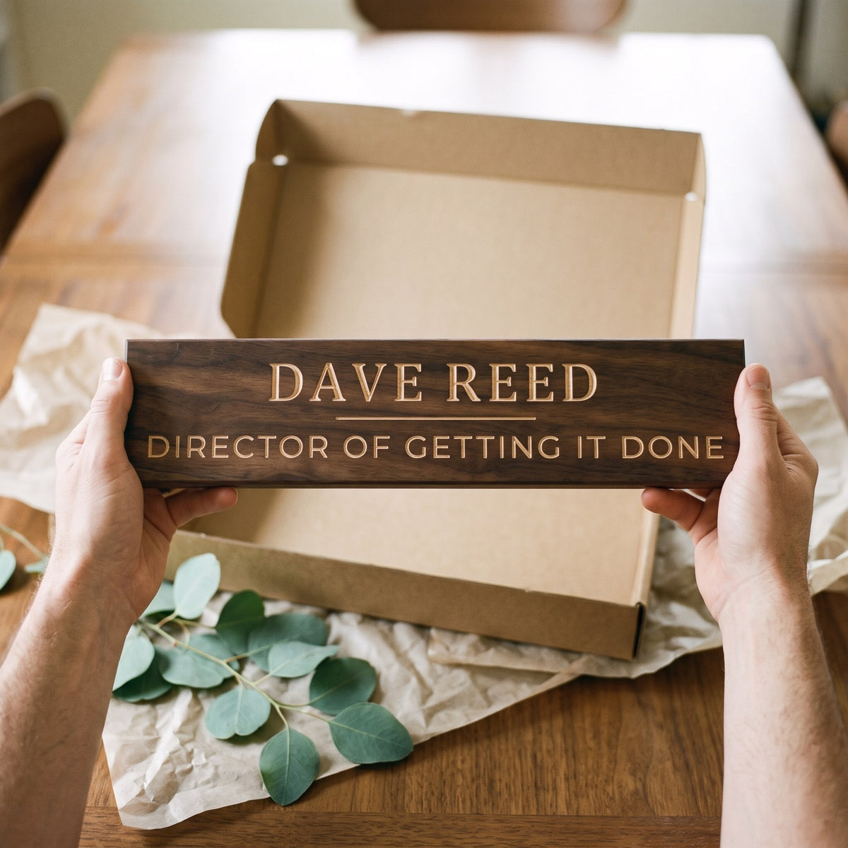 Hands holding a personalized wood sign that reads Dave Reed over a kraft gift box with tissue paper and twine
