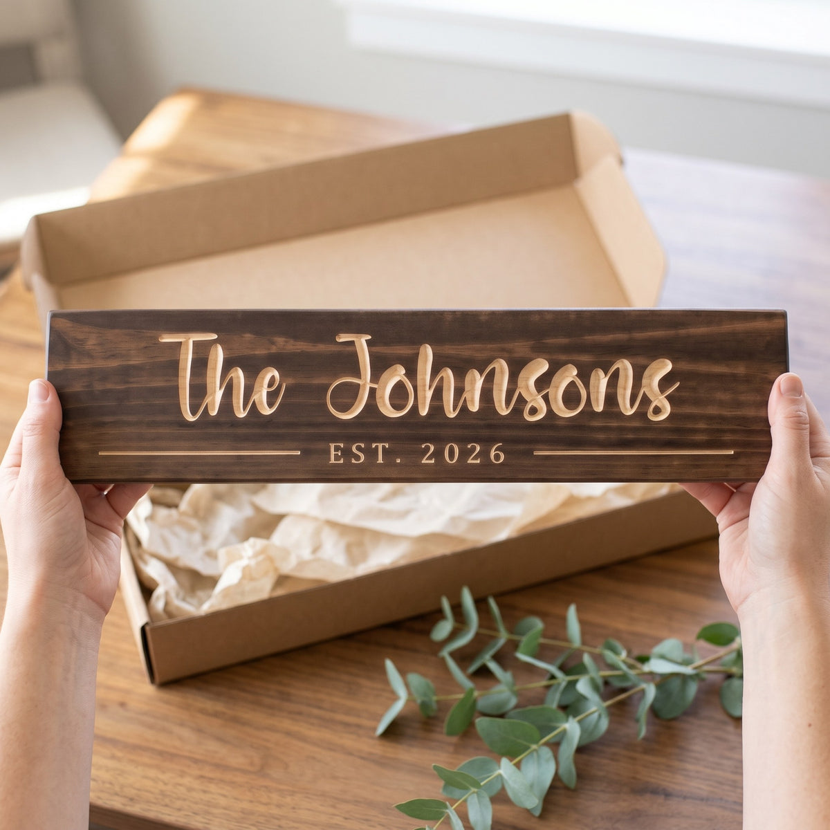 Hands holding a personalized wood sign that reads The Johnsons over a kraft gift box with tissue paper and twine