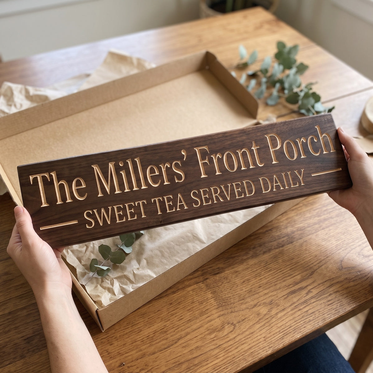 Hands holding a personalized wood sign that reads The Millers' Front Porch over a kraft gift box with tissue paper and twine