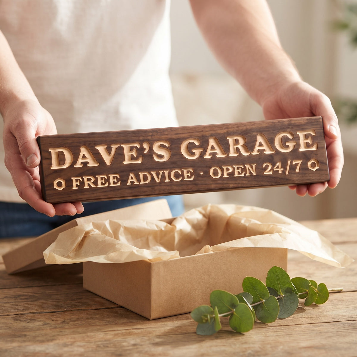 Hands holding a personalized wood sign that reads Dave's Garage over a kraft gift box with tissue paper and twine