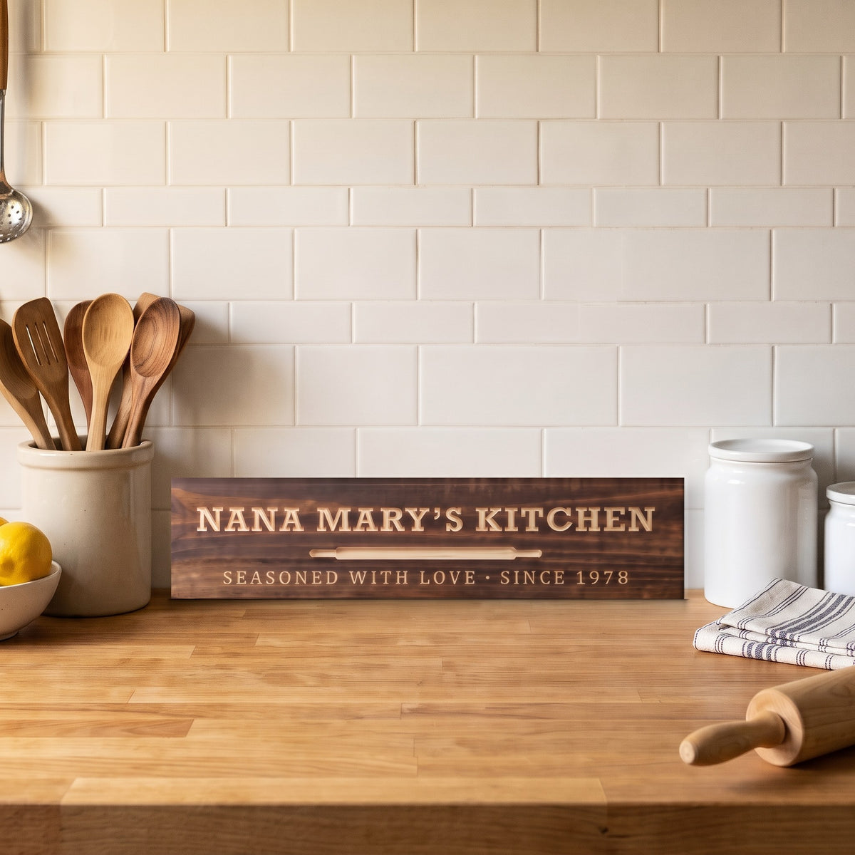 Nana Mary's Kitchen personalized wood sign standing on a butcher-block counter against subway tile beside a crock of wooden spoons, lemons and a rolling pin