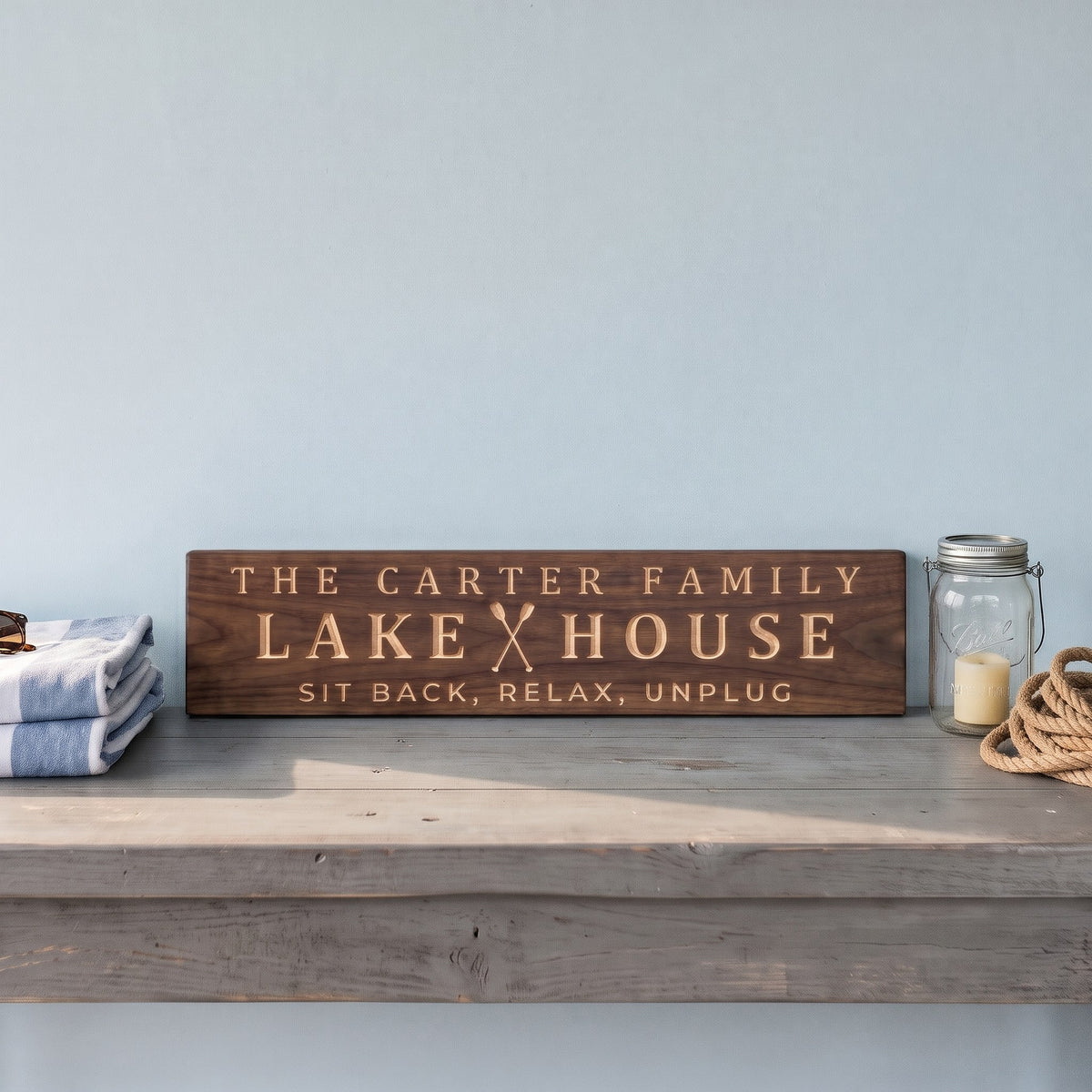 The Carter Family Lake House personalized wood sign standing on a weathered gray console table against a pale blue wall beside beach towels, a mason-jar lantern and coiled rope