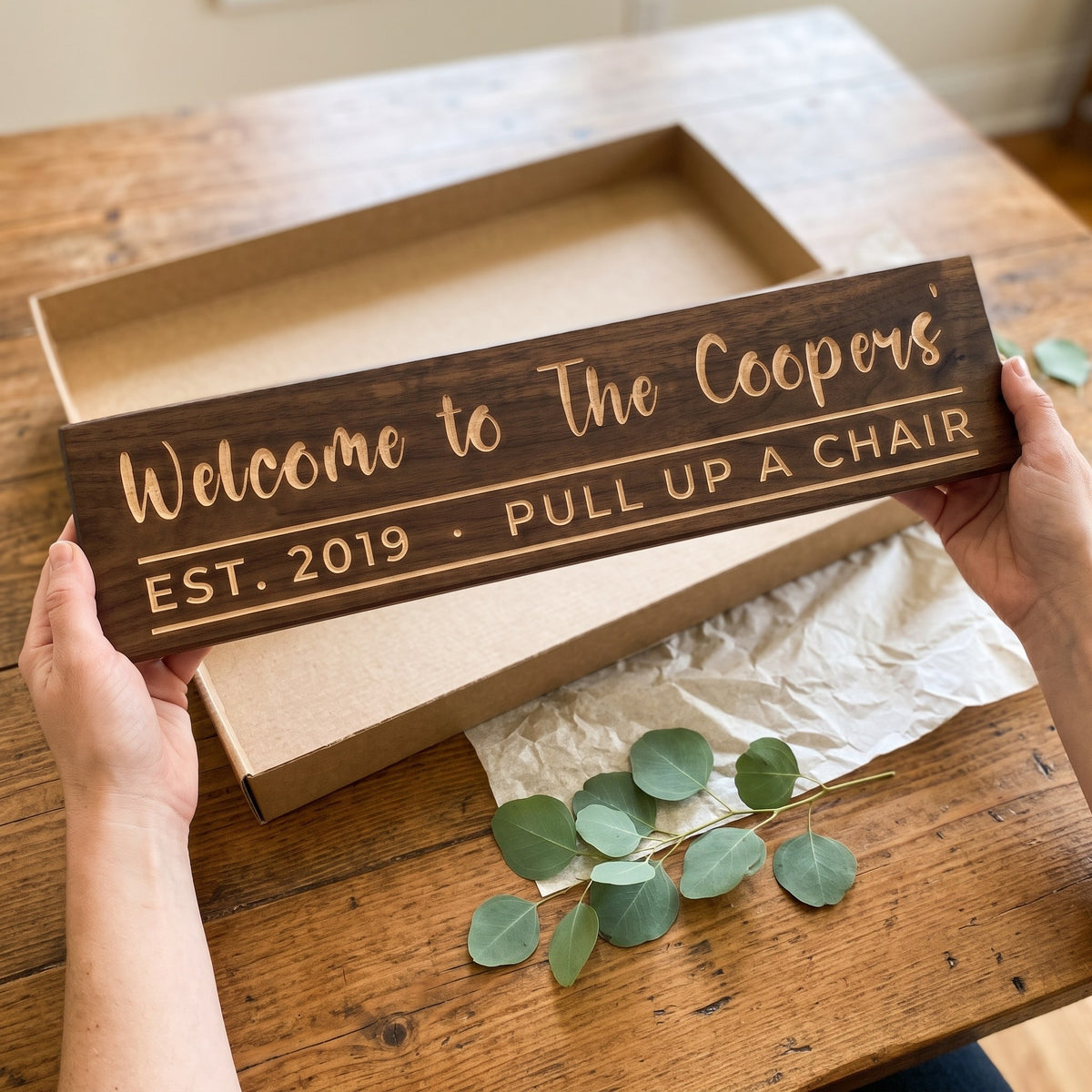 Hands holding a personalized wood sign that reads Welcome to The Coopers' over a kraft gift box with tissue paper and twine