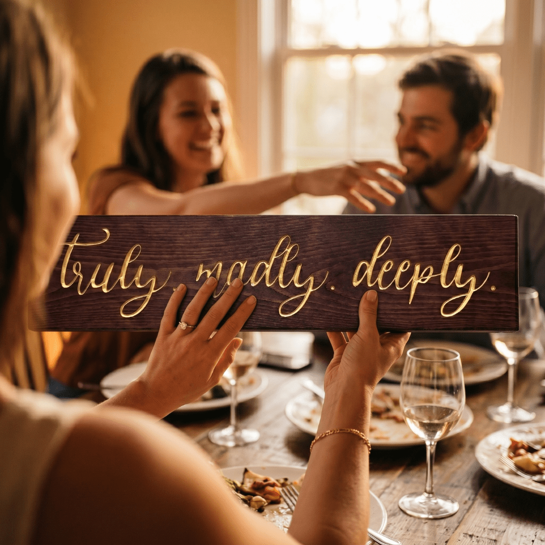 Woman with an engagement ring holding the Truly Madly Deeply carved wood sign across a dinner table as friends celebrate together with wine