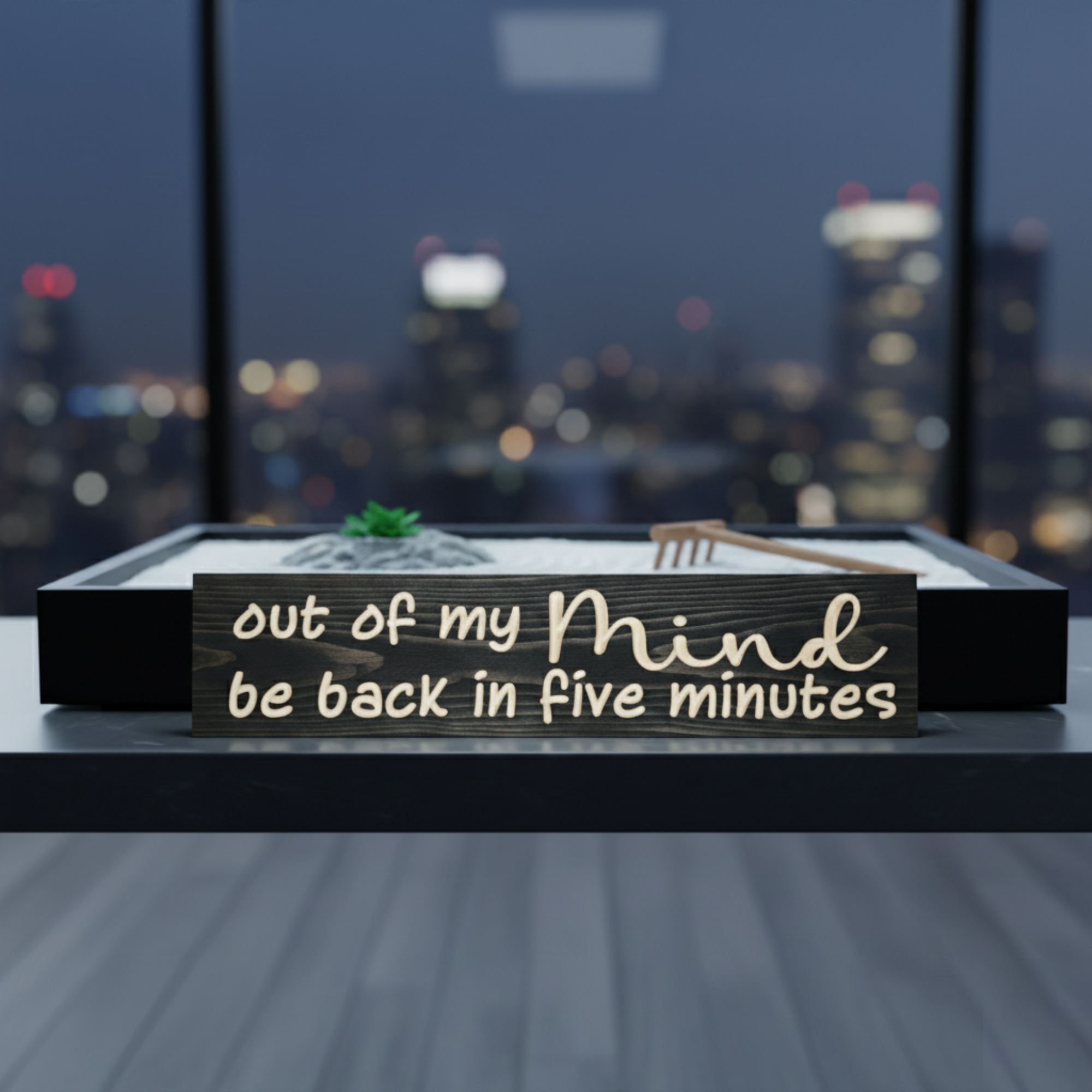 Humorous wooden desk sign "be back in five minutes" placed in front of a desktop Zen garden with a city skyline at night.