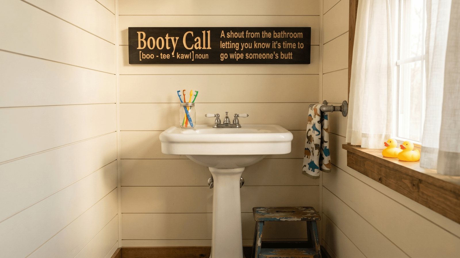 Funny hardwood Booty Call bathroom sign hanging above a white pedestal sink in a cream shiplap kids' bathroom, with rubber ducks on the windowsill, colorful toothbrushes in a mason jar, and a small wooden step stool on the floor