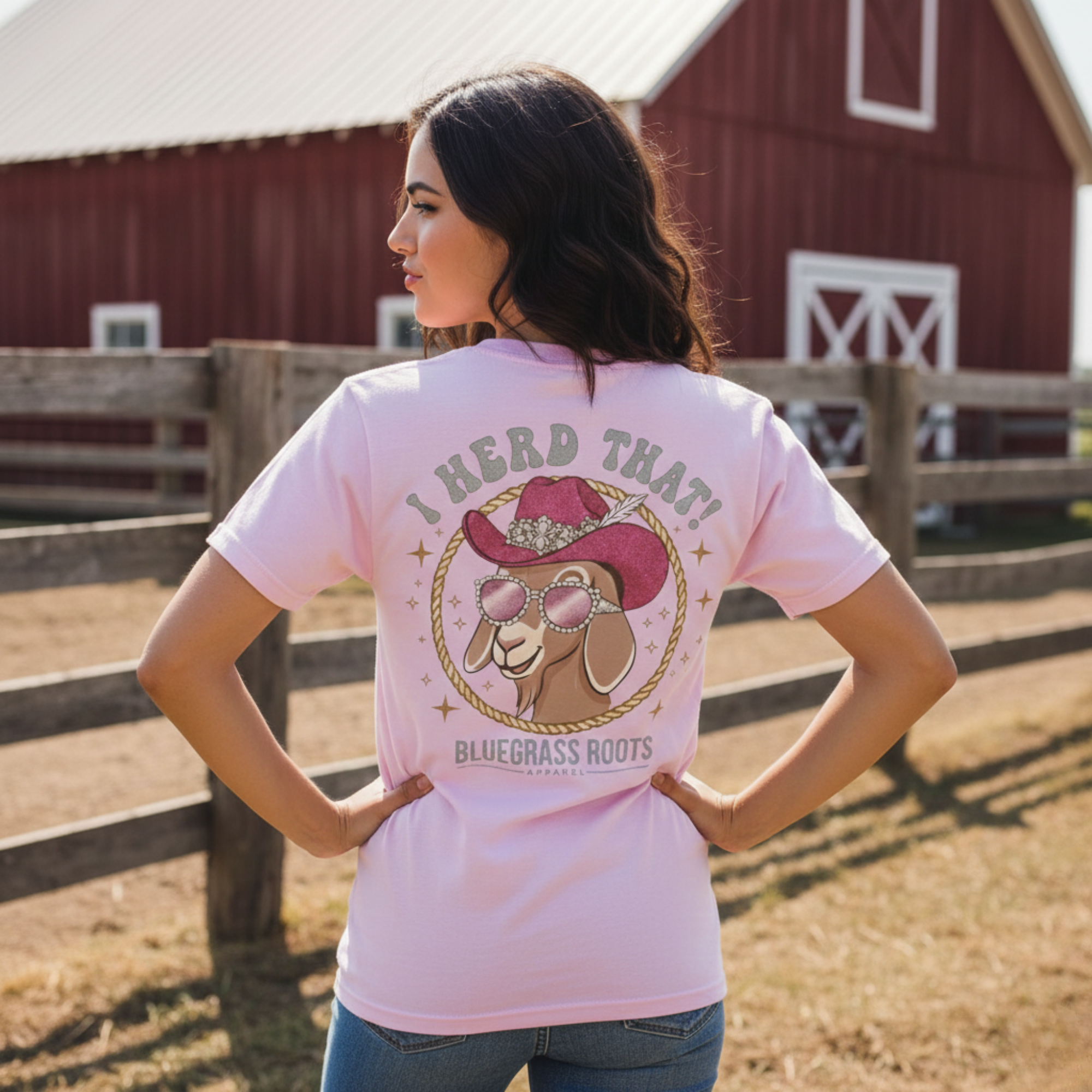 A woman wearing the pink "I Herd That" glam goat graphic tee in front of a red barn, showcasing the Western boho apparel style of Bluegrass Gifts.