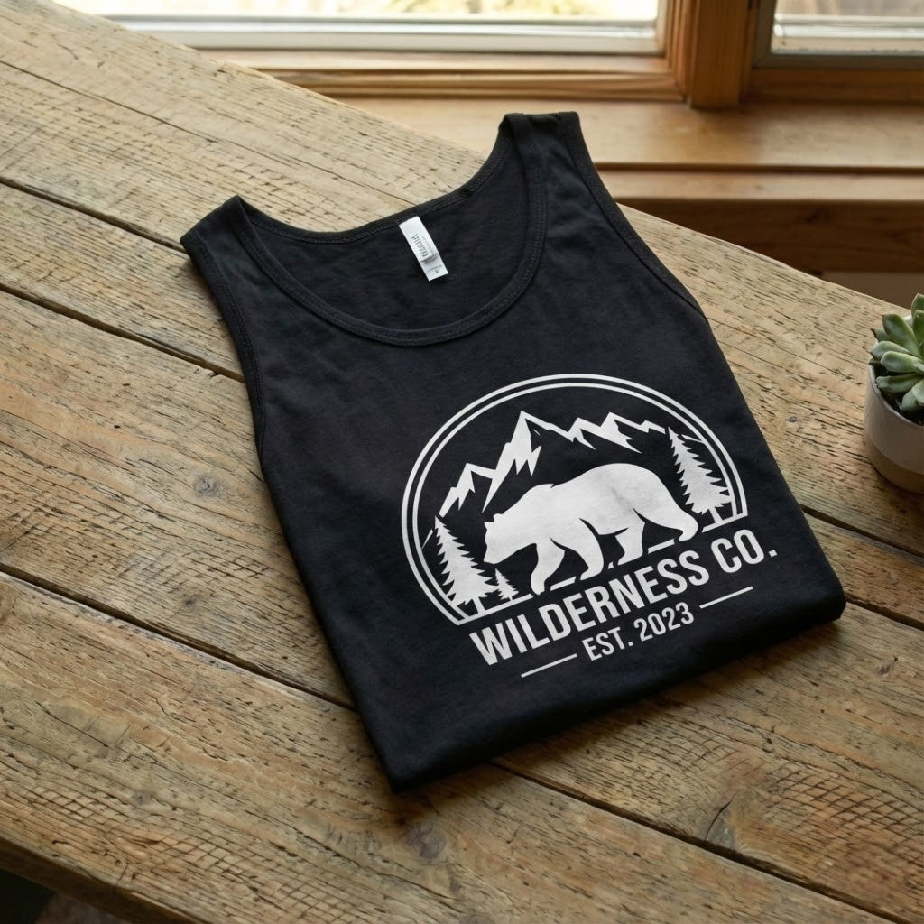 Men's black graphic tank top with white bear and mountain wilderness design folded on a rustic wooden table - Bluegrass Gifts.