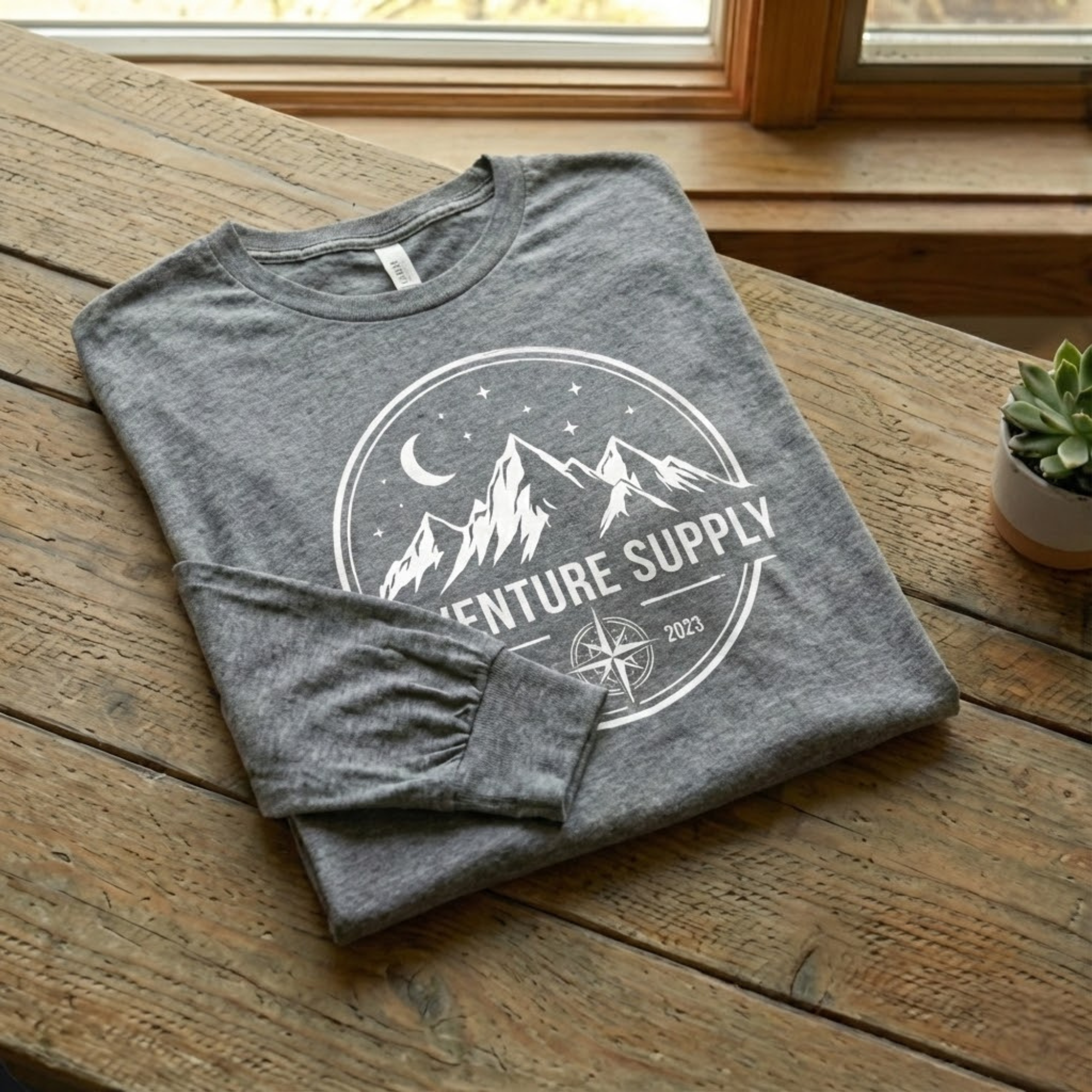 Men's grey long sleeve graphic t-shirt with white mountain and compass adventure design folded on a rustic wooden table - Bluegrass Gifts.