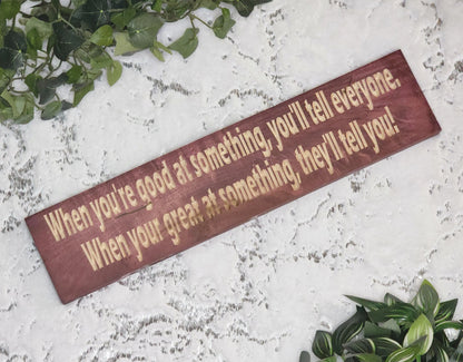 Motivational wooden sign reading When You're Good at Something, You'll Tell Everyone. When You're Great at Something, They'll Tell You! displayed in a modern home office.
