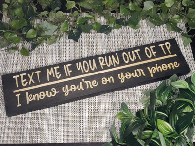 Rustic wooden bathroom sign reading Text Me if You Run Out of TP - I Know You're on Your Phone displayed in a modern guest bathroom.
