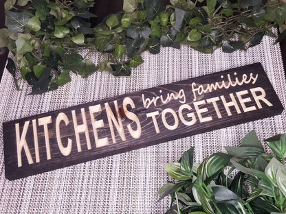Rustic wooden kitchen sign reading "Kitchens Bring Families Together" displayed in a cozy home kitchen.
