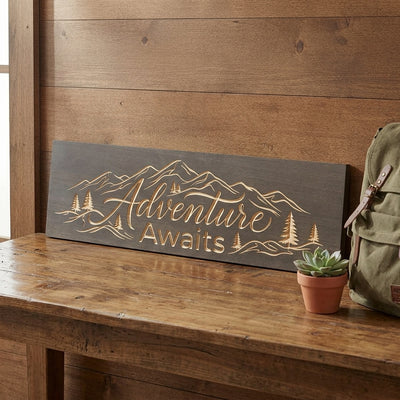 A carved wood sign with "Adventure Awaits" and mountain imagery rests on a wooden bench, a green backpack nearby, a great piece of Travel Wall Decor from Bluegrass Gifts