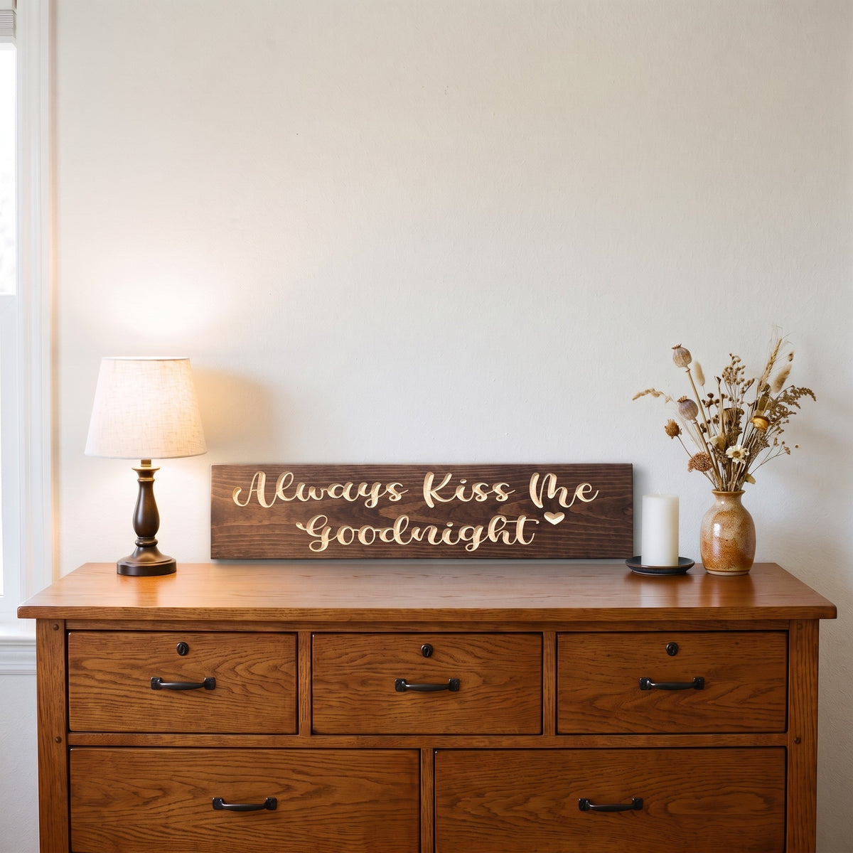 The Always Kiss Me Goodnight sign standing on a wooden bedroom dresser between a table lamp and a candle with dried flowers