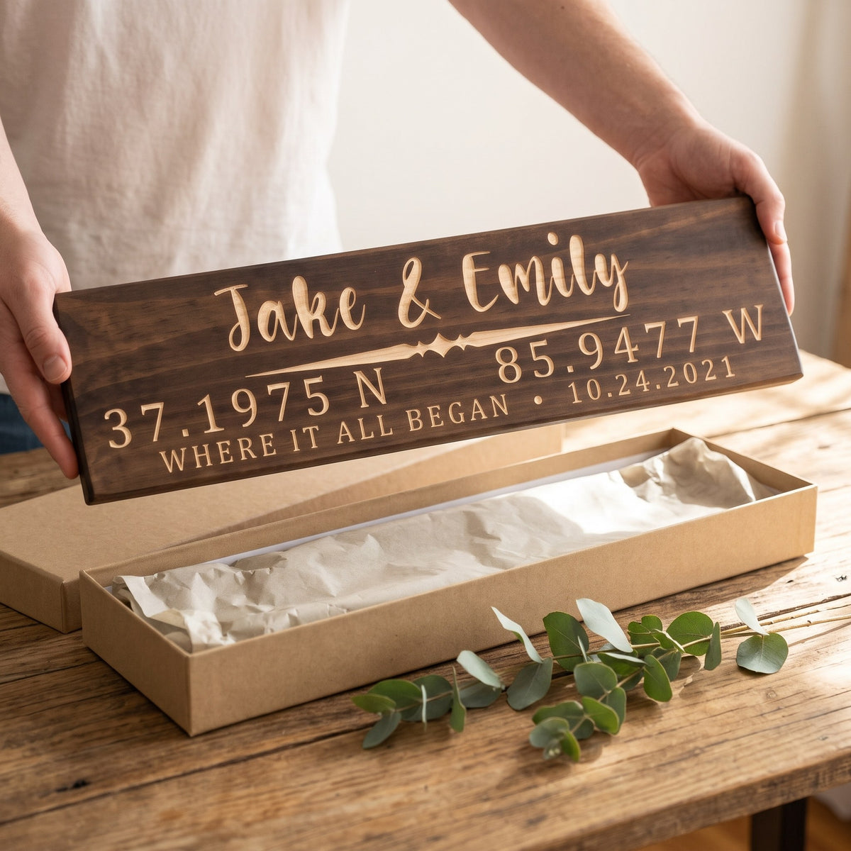 Hands holding a personalized wood sign that reads Jake & Emily over a kraft gift box with tissue paper and twine