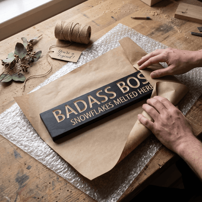 Hands wrapping a Badass Boss wood sign in kraft paper for a Boss's Day gift.