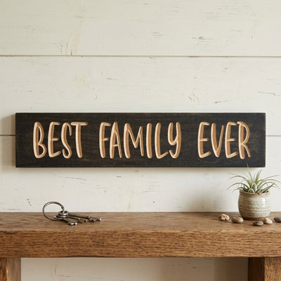 A dark wood Best Family Ever Sign hangs on a light wood wall, above a shelf with keys and a plant, from Bluegrass Gifts