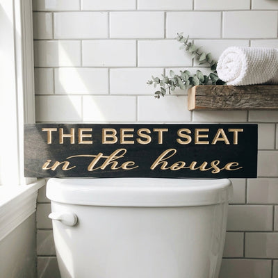 The Best Seat in the House Sign, a dark wood sign with carved light letters, rests on a white toilet in a tiled bathroom from Bluegrass Gifts