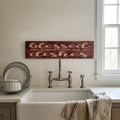 A dark reddish-brown carved wood sign, "Blessed ARE THOSE WHO DO MY dishes", mounted as Above Kitchen Sink Decor over a white farmhouse sink, from Bluegrass Gifts