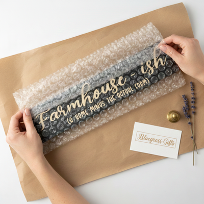 Hands carefully unboxing a Farmhouse-ish carved sign from bubble wrap and craft paper. Includes a Bluegrass Gifts business card, showcasing our secure checkout and 5-7 day shipping process.