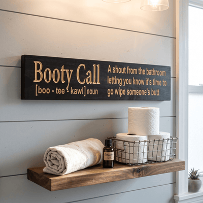 Booty Call funny bathroom sign hanging on white shiplap above a floating shelf with toilet paper, rolled towel, and essential oil bottle