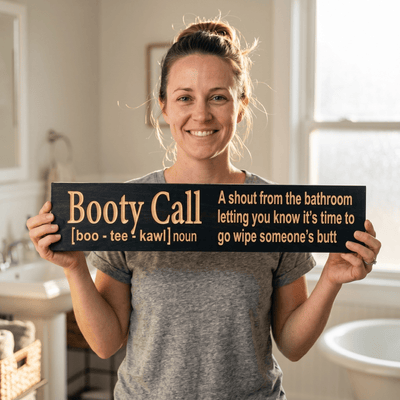 Smiling mom holding the Booty Call funny parenting bathroom sign in her bathroom, a perfect baby shower gift for new parents from Bluegrass Gifts