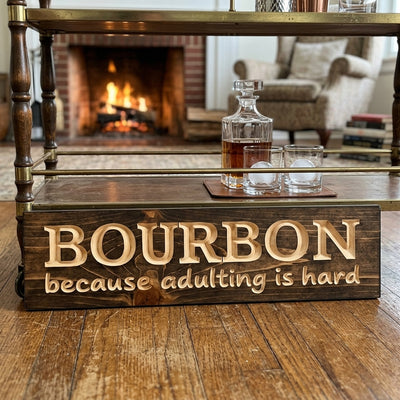 A dark wood Adulting Sign with carved letters sits on a bar cart next to a bourbon decanter and glasses, with a fireplace in the background from Bluegrass Gifts