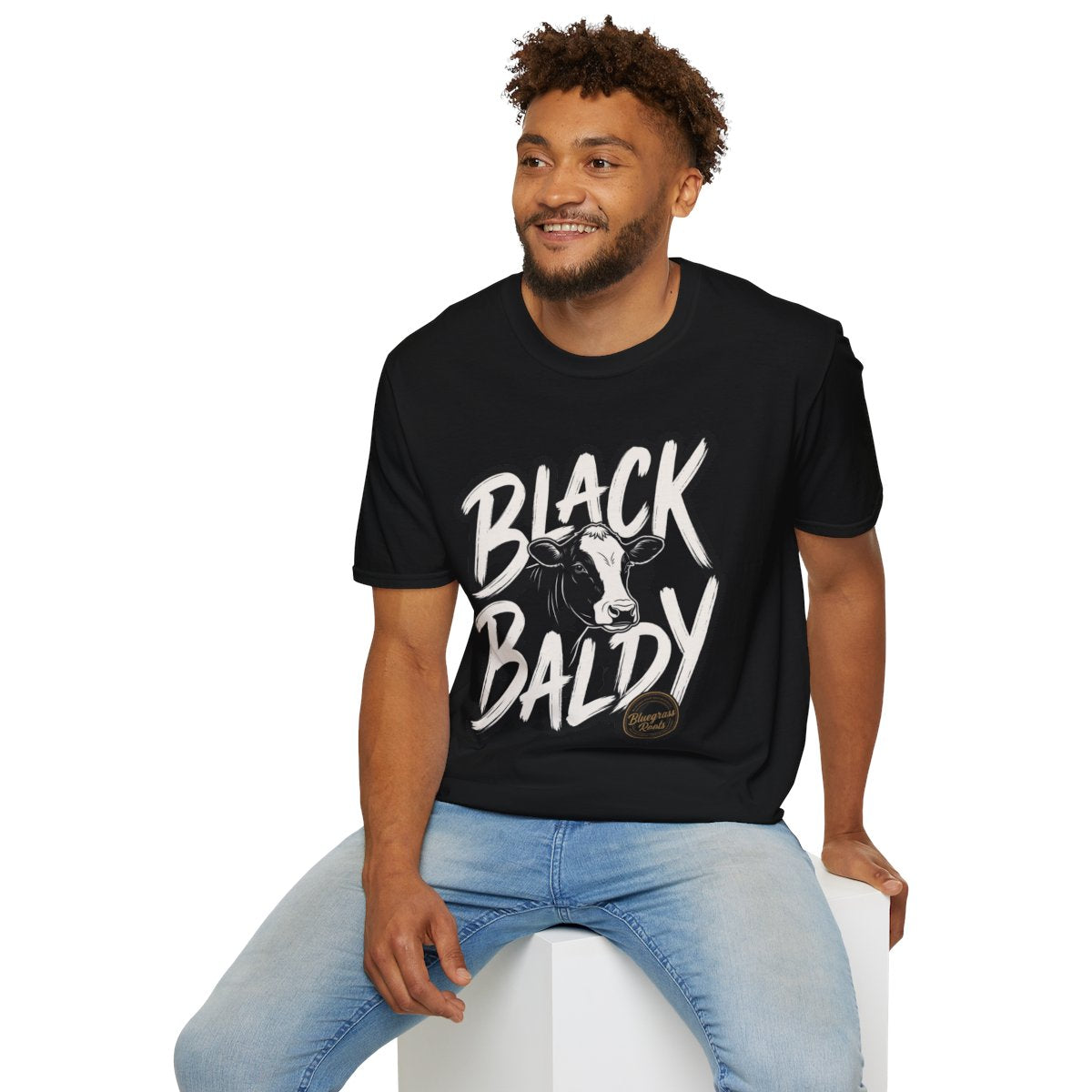 A man sitting comfortably on a white cube, modeling the black Cattle Shirt - Black Baldy with its full design