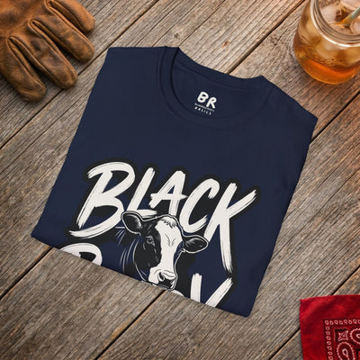 A navy blue "Cattle Shirt - Black Baldy" tee folded on a rustic wooden surface, next to a leather glove and drink