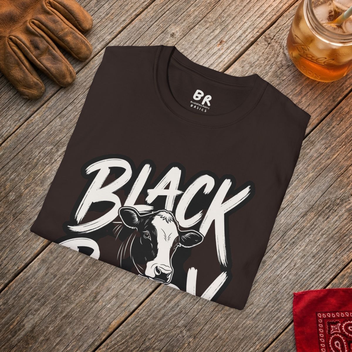 A dark brown Cattle Shirt - Black Baldy folded on a rustic wooden surface with a glove and iced tea