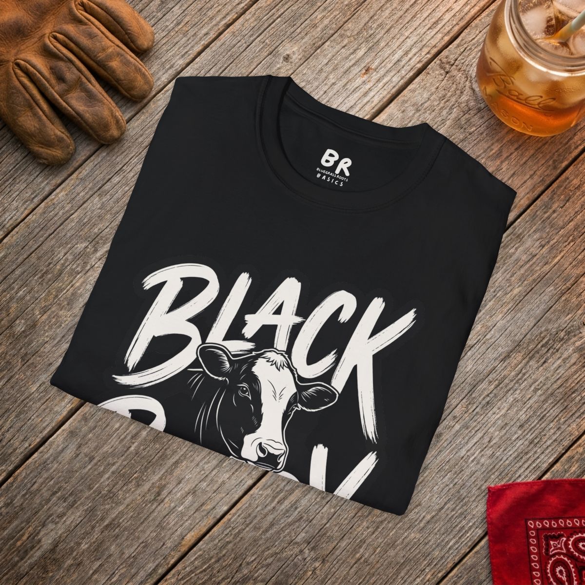 A black Cattle Shirt - Black Baldy folded on a rustic wooden surface, with a work glove and iced tea nearby