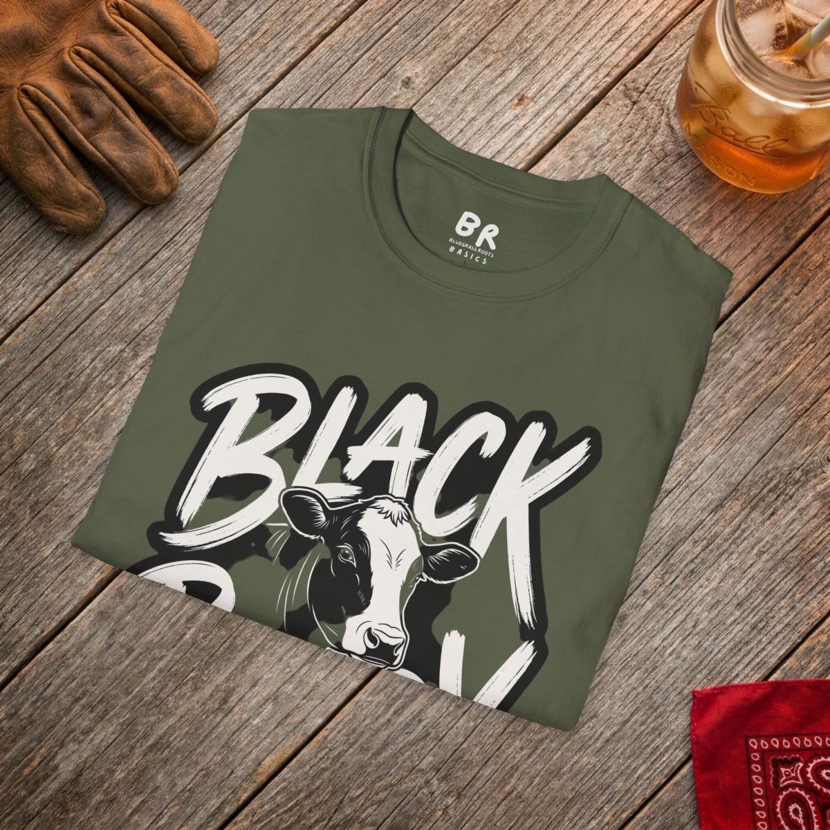 An olive green "Cattle Shirt - Black Baldy" tee folded on a rustic wooden surface, next to a leather glove and drink