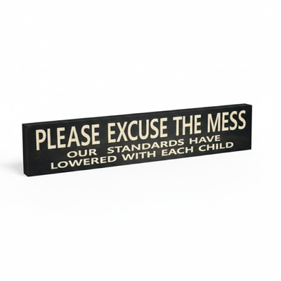 Angled view of a deep-carved wooden home decor sign with the funny parenting quote "Please Excuse the Mess Our Standards Have Lowered With Each Child".