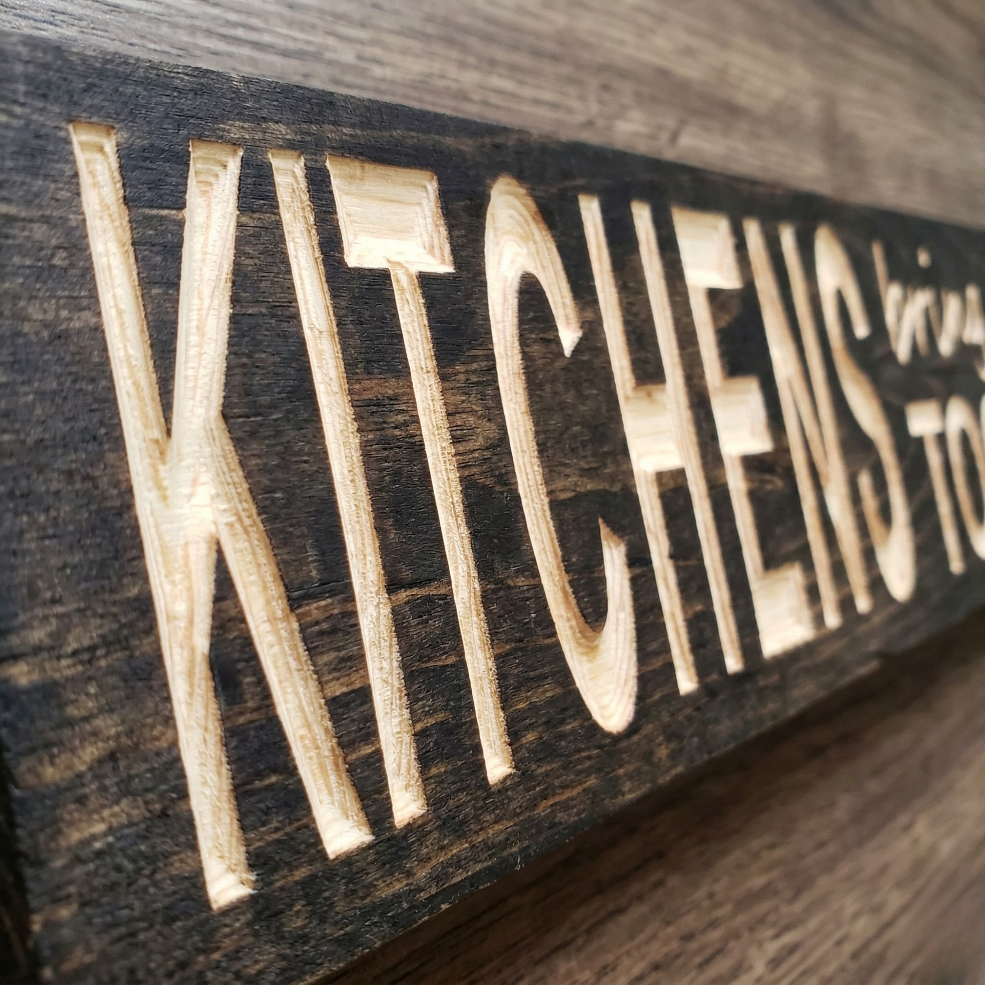 Close-up detail of the deep CNC carving on a solid wood sign, showing the natural grain within the letters of the word "KITCHENS