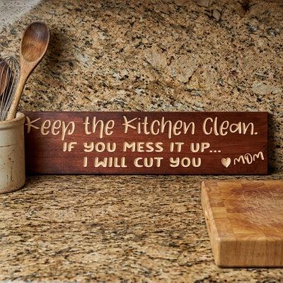 A close-up of the carved Cooking Sign resting on a speckled granite kitchen counter next to kitchen utensils from Bluegrass Gifts