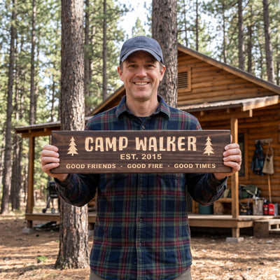 A smiling man in a flannel shirt and a plain ball cap standing outside a log cabin among tall pines holds a personalized wood sign that reads Camp Walker