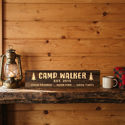 Camp Walker personalized wood sign standing on a rustic log mantel against a knotty pine wall beside a lantern, a camp mug and a plaid blanket