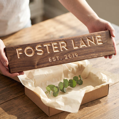 Hands holding a personalized wood sign that reads Foster Lane over a kraft gift box with tissue paper and twine